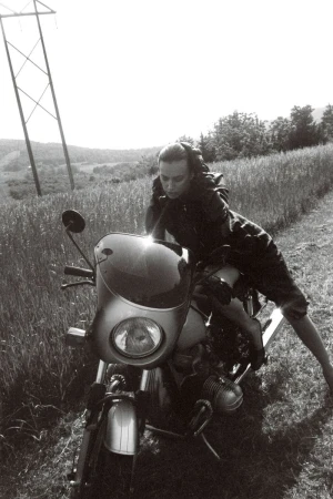 Monochrome shot of a woman in casualwear posing on a vintage motorcycle on a grassy path with power lines in the background.
