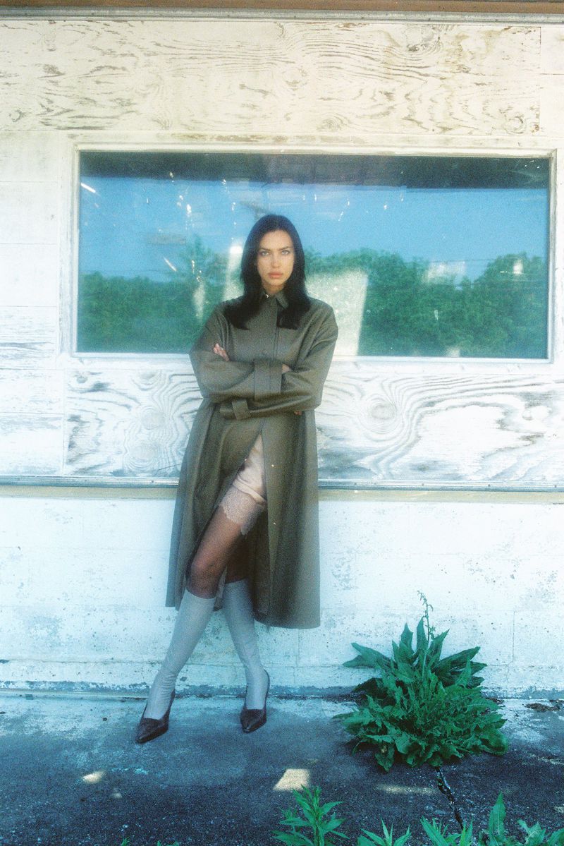 An adult female model poses in a long belted coat, sheer hosiery, and heels outside of a building with a boarded window.