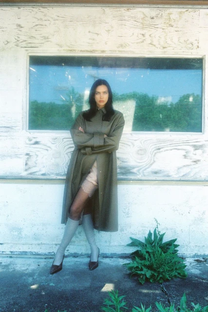 An adult female model poses in a long belted coat, sheer hosiery, and heels outside of a building with a boarded window.