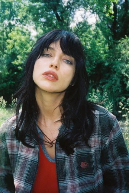 Portrait of a woman with dark hair and bangs, wearing a plaid jacket, outdoors in a natural setting with green foliage and trees.