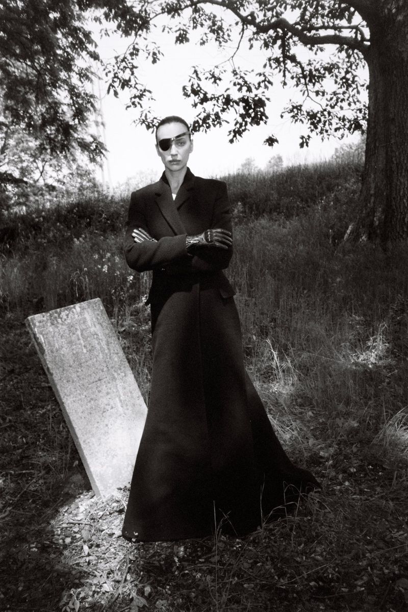 Woman in a long black coat with an eye patch stands near a gravestone in a grassy field with trees in a black and white photograph.