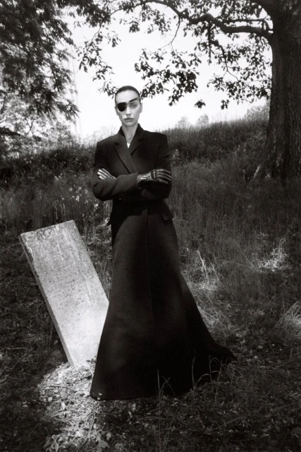 Woman in a long black coat with an eye patch stands near a gravestone in a grassy field with trees in a black and white photograph.