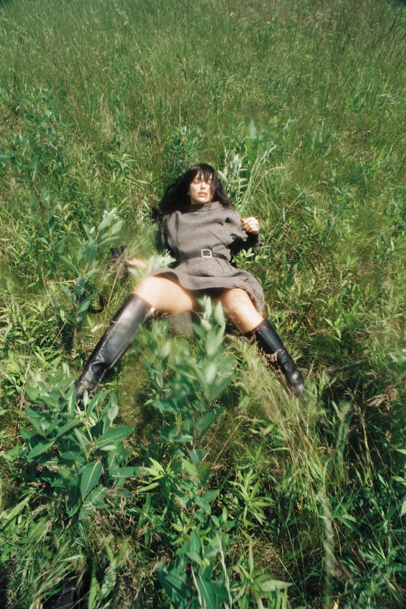 Woman lies in green field wearing vintage dress and black boots in outdoor setting, captured in high-angle shot. Tall grass surrounds the subject.