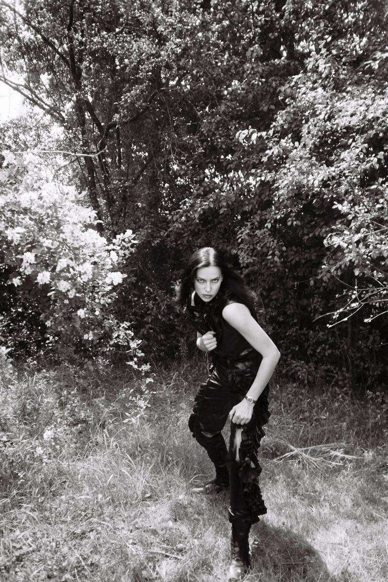 A woman in dark clothing poses in an outdoor setting with trees. The image is monochrome and reminiscent of a vintage photograph.
