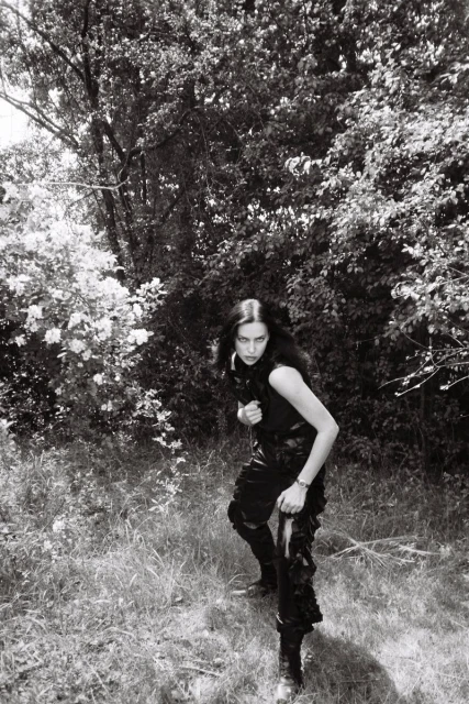 A woman in dark clothing poses in an outdoor setting with trees. The image is monochrome and reminiscent of a vintage photograph.