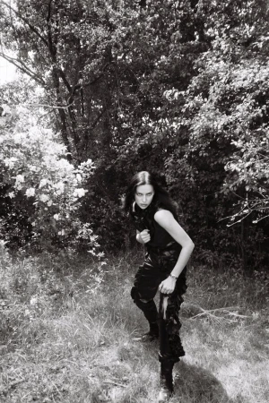 A woman in dark clothing poses in an outdoor setting with trees. The image is monochrome and reminiscent of a vintage photograph.