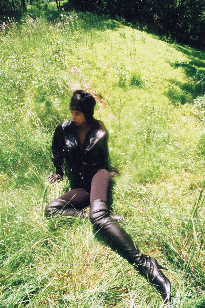 Woman poses sitting in tall grass wearing a black jacket and tall leather boots in an outdoor setting with dappled light.