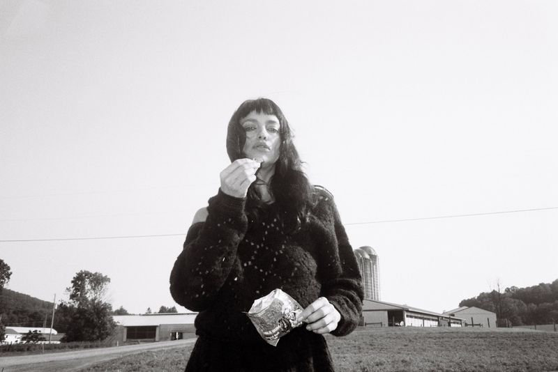 A woman in a black sweater stands in a rural field and eats potato chips in a black and white photograph with a farmhouse in background.