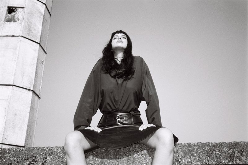 Low angle monochrome shot of model Irina Shayk seated on a concrete ledge wearing a dark belted top against an open sky.