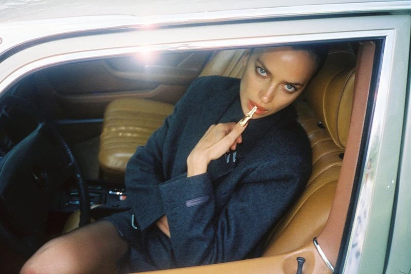 Irina Shayk, in a grey jacket, poses inside a car with tan leather interior, holding a gold makeup applicator to her lips.