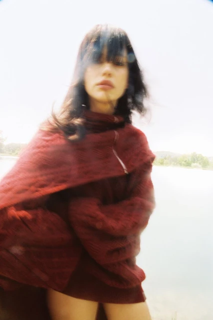 Woman in stylish maroon sweater poses near a lake, creating a soft, dreamy portrait with diffused natural lighting and serene background.