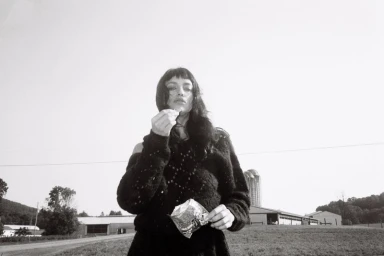Irina Shayk wearing a dark textured sweater holding a small snack package outdoors with distant farm buildings