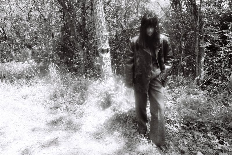 Model Irina Shayk wearing a dark leather jacket and wide-leg trousers standing in a wooded forest in black and white.