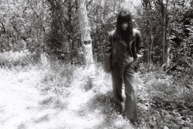 Model Irina Shayk wearing a dark leather jacket and wide-leg trousers standing in a wooded forest in black and white.