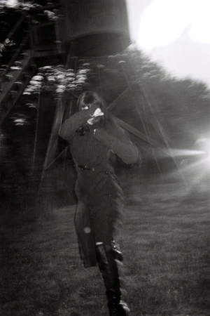 Irina Shayk running across a dark grassy field shielding her face, wearing a dark knit dress and tall black boots in low light.