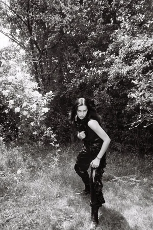 Irina Shayk crouching in a dark sleeveless top and ruched trousers amidst outdoor woodland foliage in black and white.