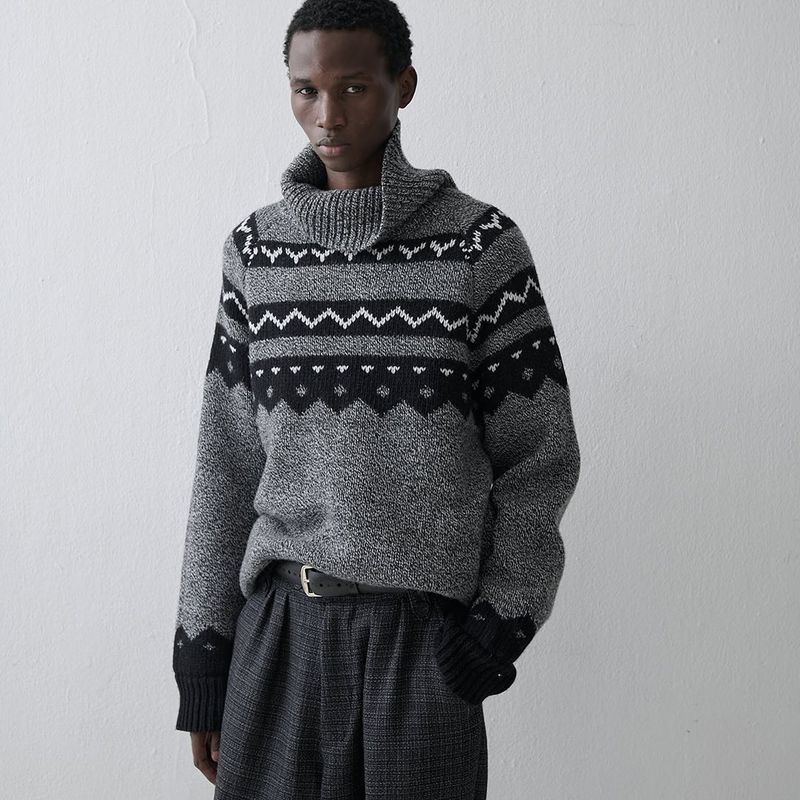 Young man in gray patterned turtleneck sweater and pleated pants poses indoors against a white wall in a fashion shot.