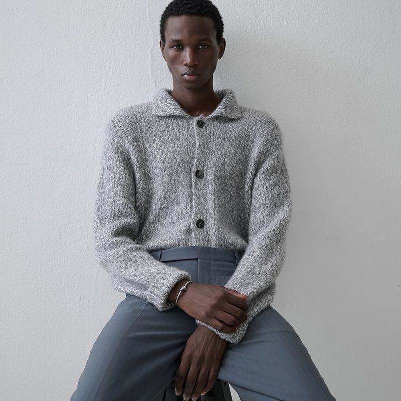 Close-up studio shot of a Black male model wearing a grey collared sweater and blue pants, sitting against a white wall, looking at the camera.