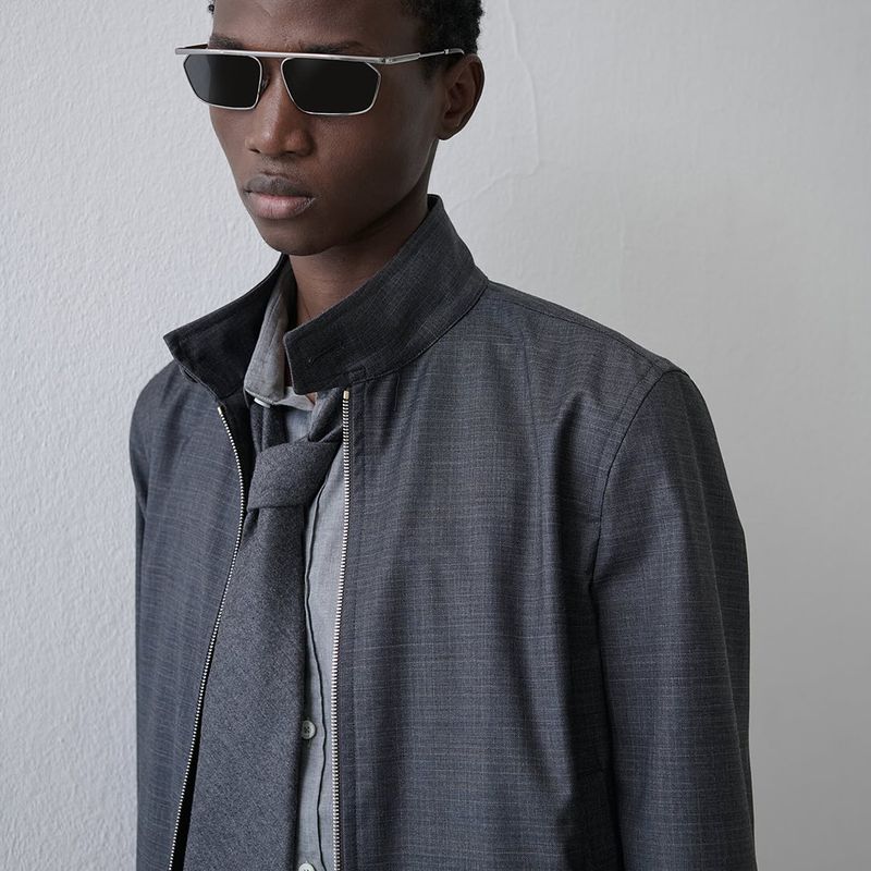 Studio portrait of a young Black man wearing sunglasses, grey collared jacket, and tie against a neutral background.