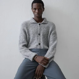 Model Gaye Serigne seated wearing a textured grey knit button-down cardigan and grey trousers against a plain light wall.