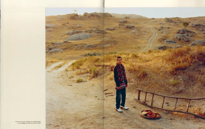 Young man stands on a dusty path in an arid landscape with dry grasses, rocks, and hills under a soft light. Ladder and basket nearby.