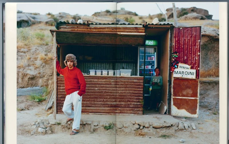 Two people stand outside a small, rustic shop in a rocky, desert landscape. A refrigerator, snacks, and hand-painted signage are visible.