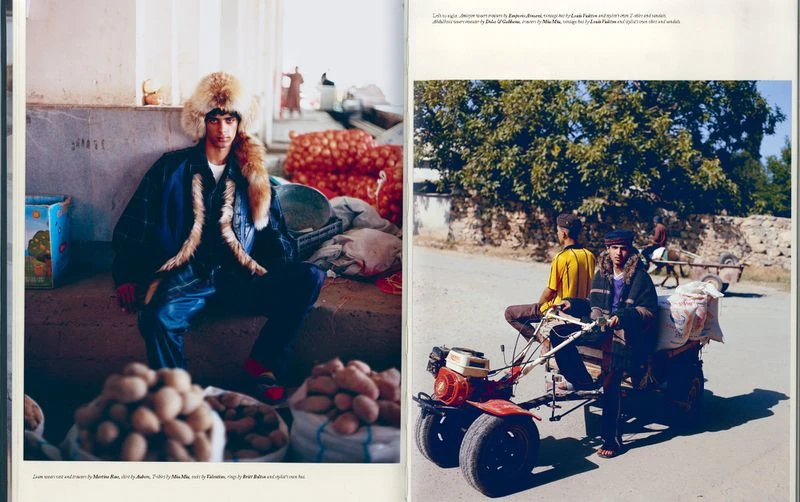 Outdoor fashion shot with two men, one in fur hat and jacket, the other seated on a tractor-pulled cart, set in a village.