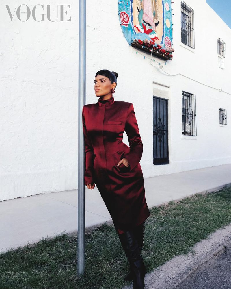 America Ferrera wears a structured burgundy satin coat dress while leaning on a pole in front of a white building for Vogue Mexico.