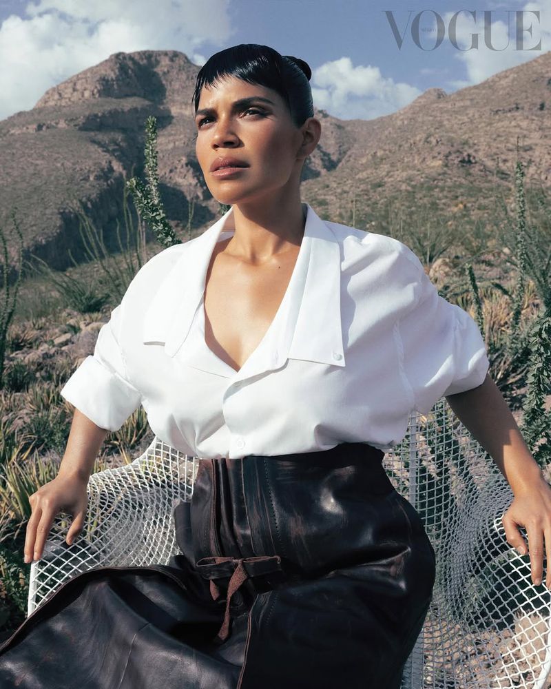 Adult woman poses in a white collared shirt and dark leather skirt in the desert, with mountains and desert plants in the background.