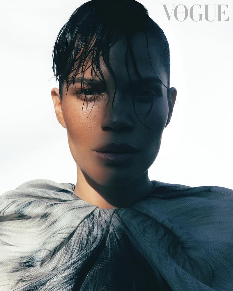 Glamorous portrait of an adult female with wet dark hair wearing a gray feathered collar against a bright white backdrop. Vogue magazine.