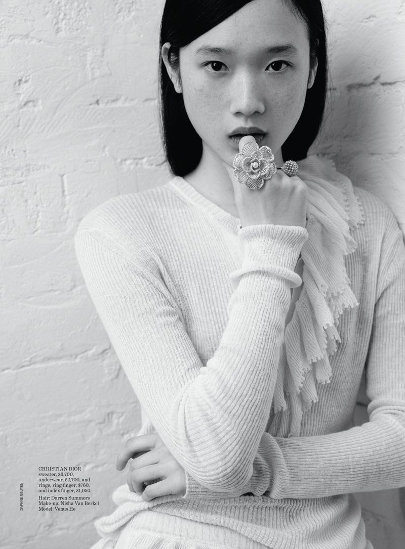 Black and white portrait of a young Asian woman in a ribbed sweater and large floral ring, posing against a white brick wall.