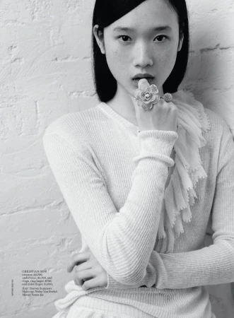 Black and white portrait of a young Asian woman in a ribbed sweater and large floral ring, posing against a white brick wall.
