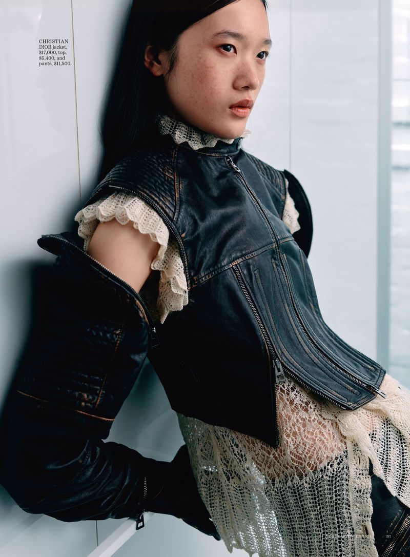 Asian woman in a leather vest and lace dress poses against a white wall with natural light. Fashion editorial shot with neutral colors.