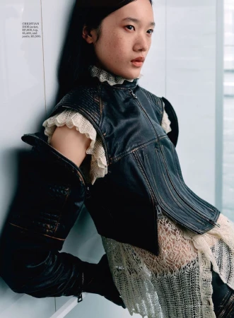 Asian woman in a leather vest and lace dress poses against a white wall with natural light. Fashion editorial shot with neutral colors.