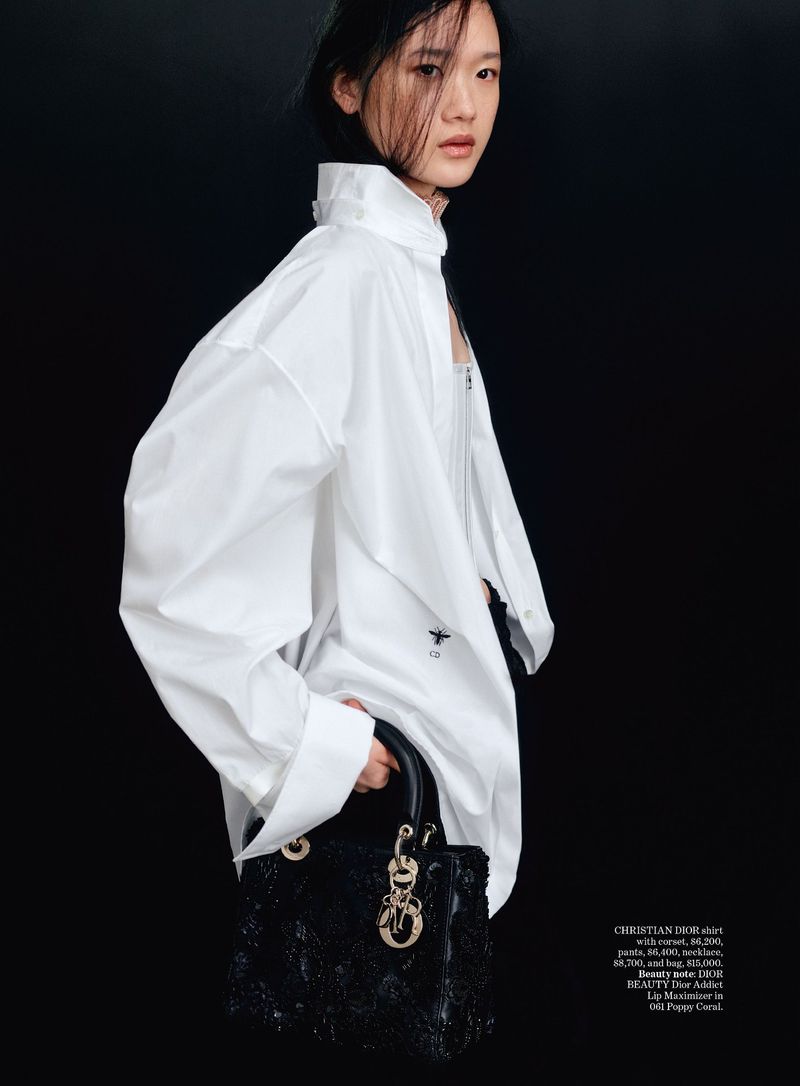 Woman poses in oversized white shirt, holding black Lady Dior handbag against dark backdrop in studio fashion editorial shot.