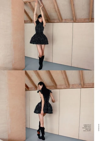 A fashionable young woman in a black dress poses indoors, complemented by black boots, against a backdrop of neutral-toned walls and wood accents.