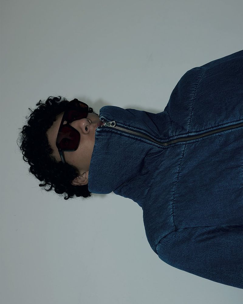 Person with dark curly hair wearing a zipped, high-necked blue denim jacket and red sunglasses, posing against a white background.