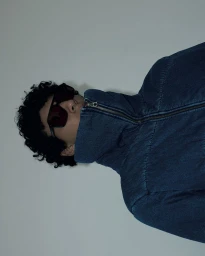 Model wearing dark sunglasses and a high-collar blue denim zip-up jacket against a neutral studio backdrop.
