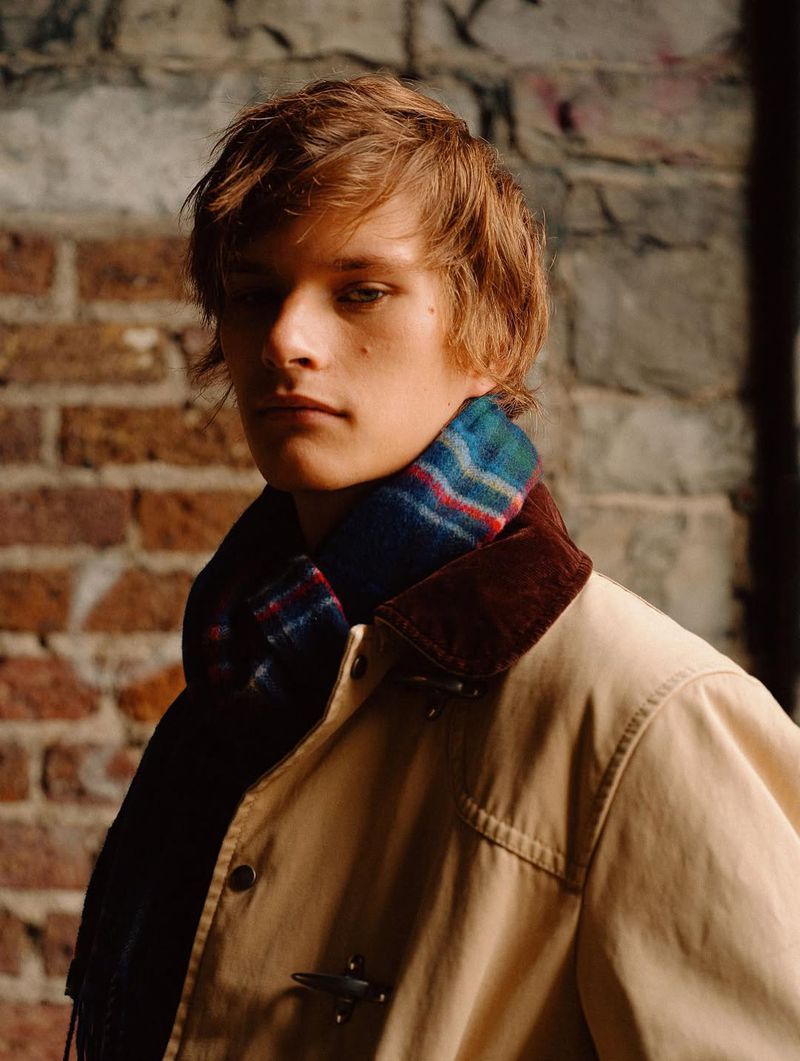 Close-up of a male model wearing a colorful scarf and a light brown coat with a brick wall as a blurred background.