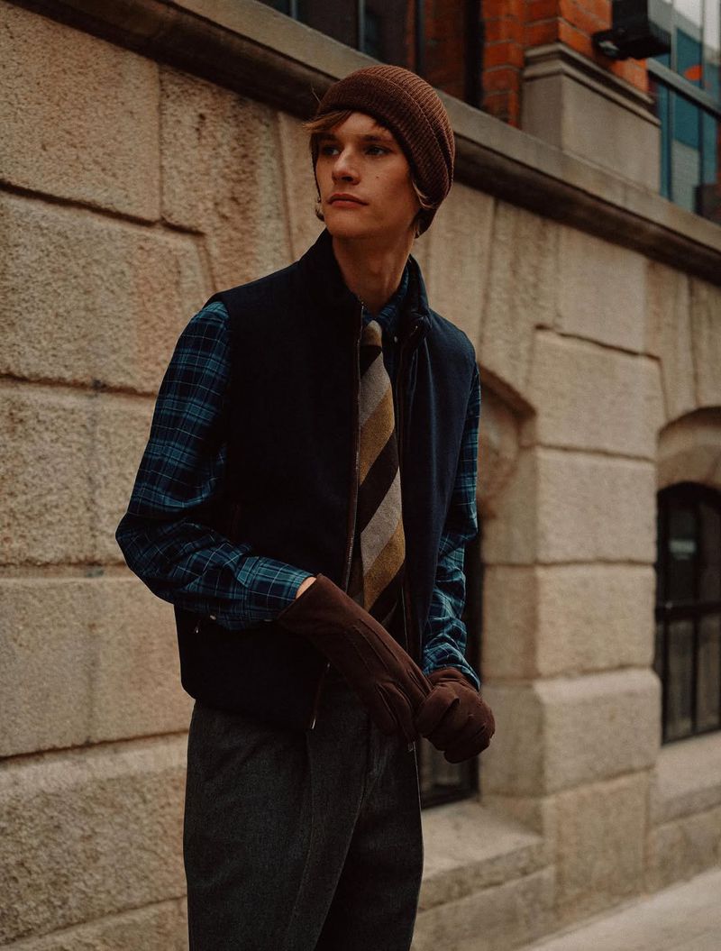 A young man in a beanie, vest, and patterned shirt poses outdoors against a brick building in an urban setting with natural light.