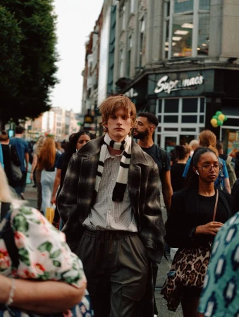 Fashionable young man in coat and scarf walking on city street surrounded by diverse pedestrians and urban buildings in daylight.