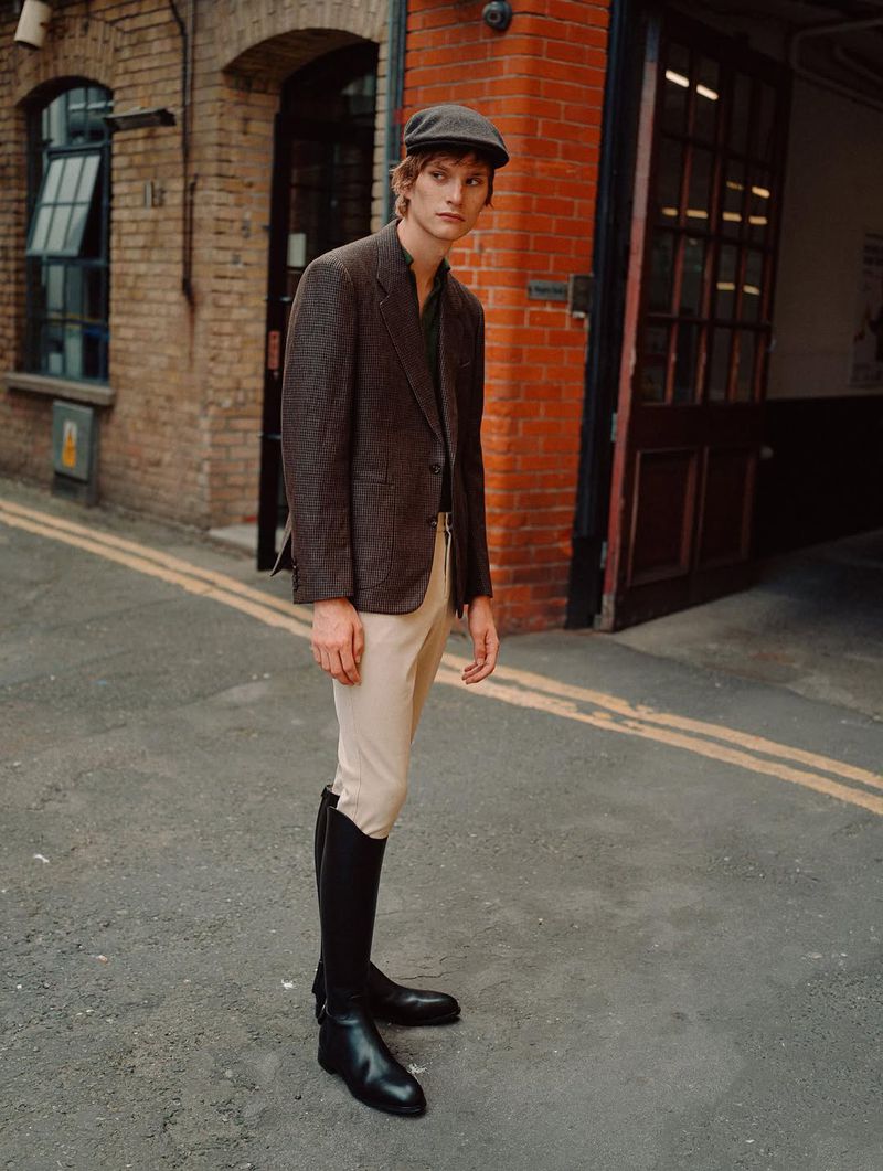 Man in tweed blazer, newsboy cap, tan pants, and tall black boots stands in urban alleyway, brick buildings in the background.
