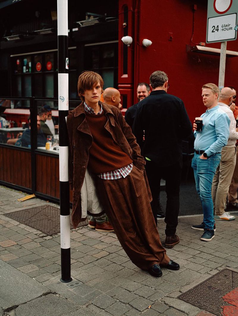 Stylish man in brown corduroy suit leans against a black and white pole outdoors, with a group of people in the background.