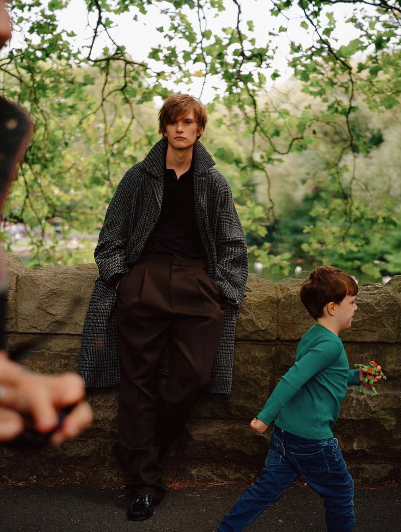 A young man in a coat poses in an outdoor setting, with a child walking by and someone partially visible holding a device.