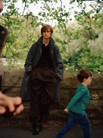 A young man in a coat poses in an outdoor setting, with a child walking by and someone partially visible holding a device.