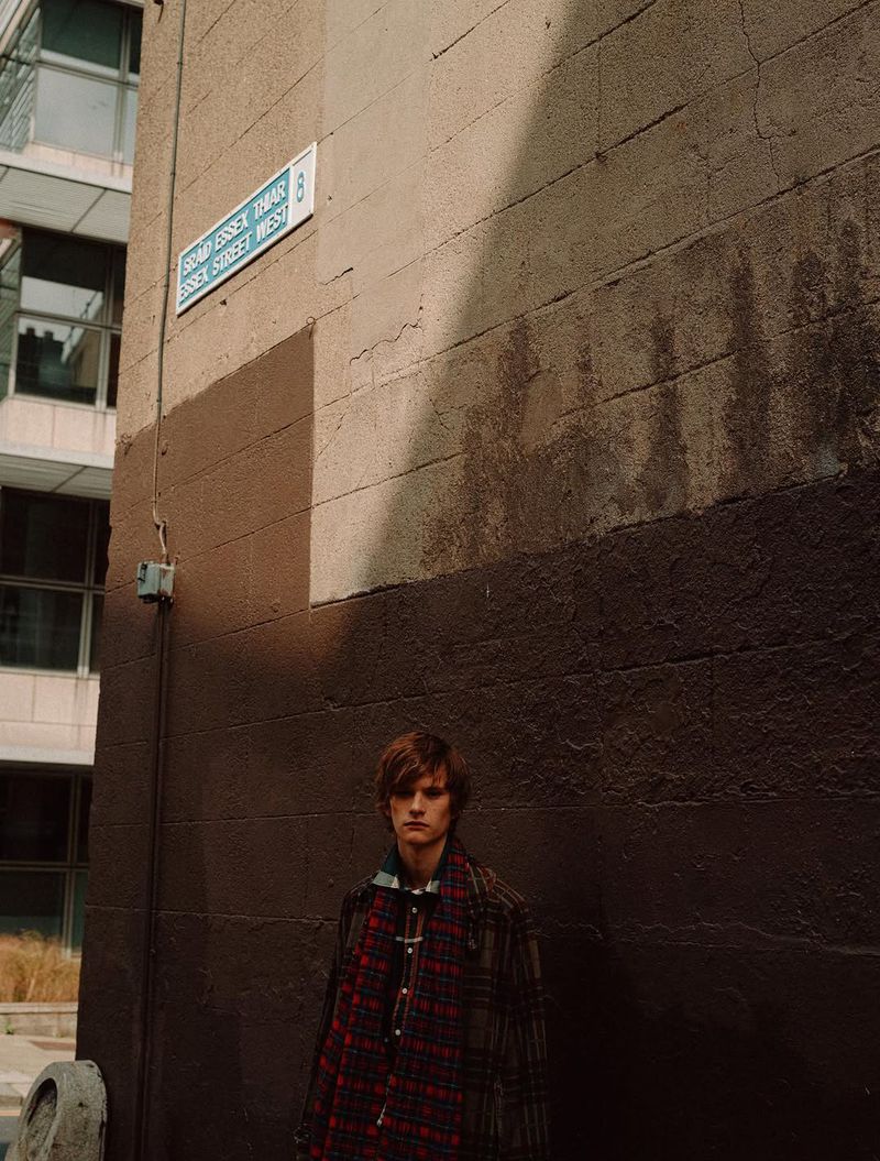 Man in a plaid coat stands in an alleyway with a street sign on a brown wall in natural light, creating a candid, urban fashion image.