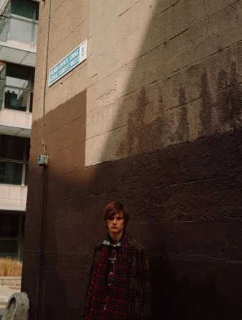 Man in a plaid coat stands in an alleyway with a street sign on a brown wall in natural light, creating a candid, urban fashion image.