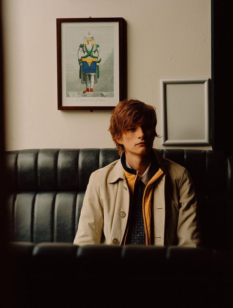 Portrait of a young adult male with auburn hair wearing a tan jacket seated on a leather bench in a dimly lit indoor setting.