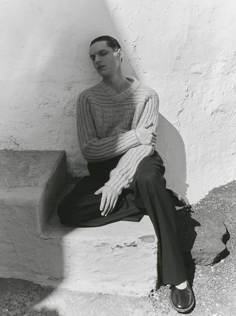 Monochrome portrait of a man wearing a textured sweater and trousers, seated on stone steps in sunlight, captured in a lifestyle setting.