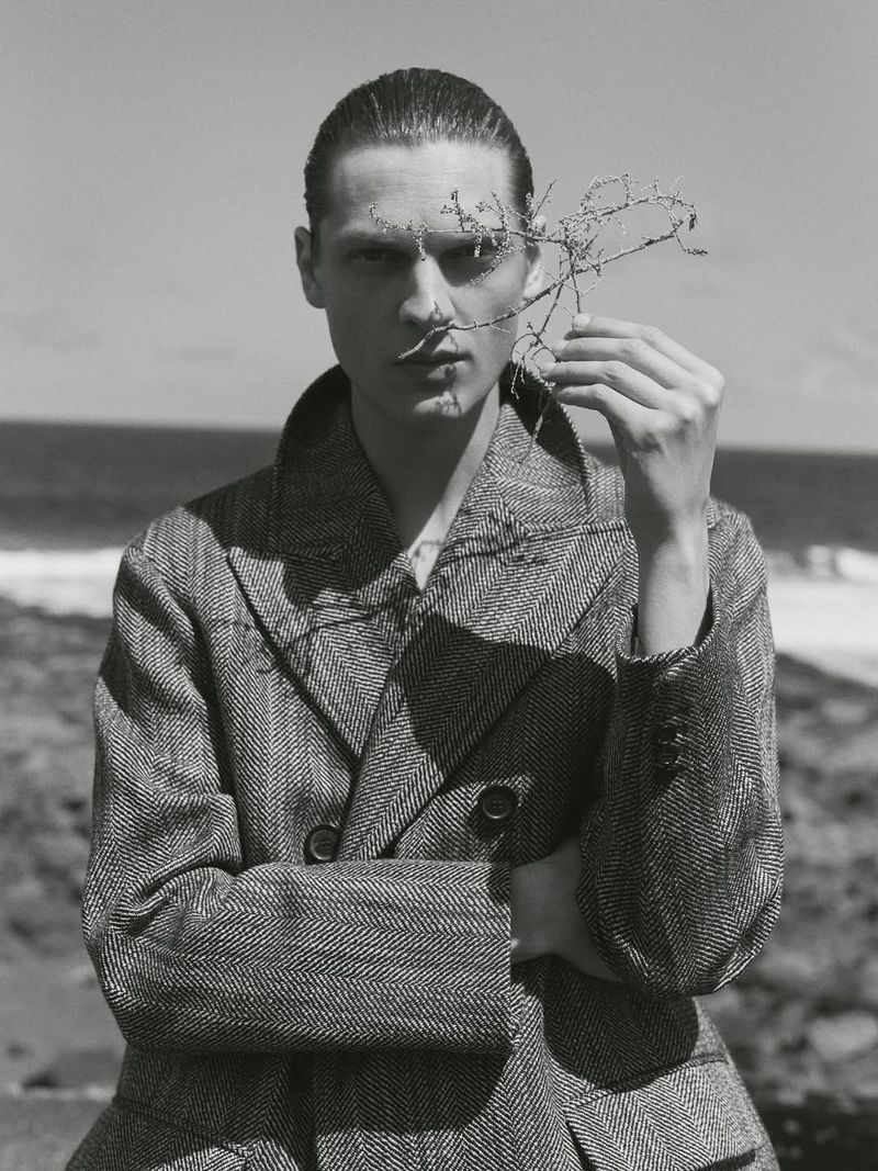 Monochrome portrait of a person in a patterned coat holding a spiky branch against a seascape backdrop, with a desaturated, artistic aesthetic.